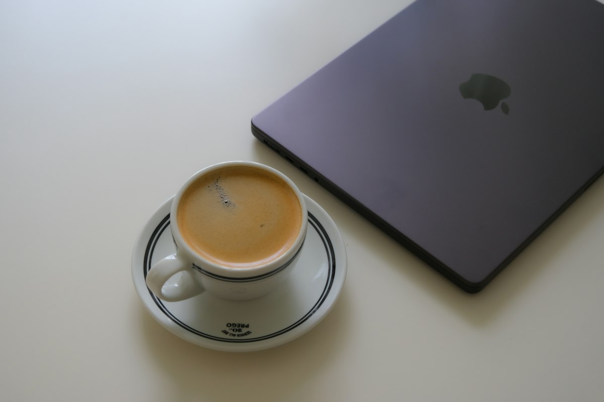 An espresso cup beside a closed laptop on a cafe table, with a notebook and morning light