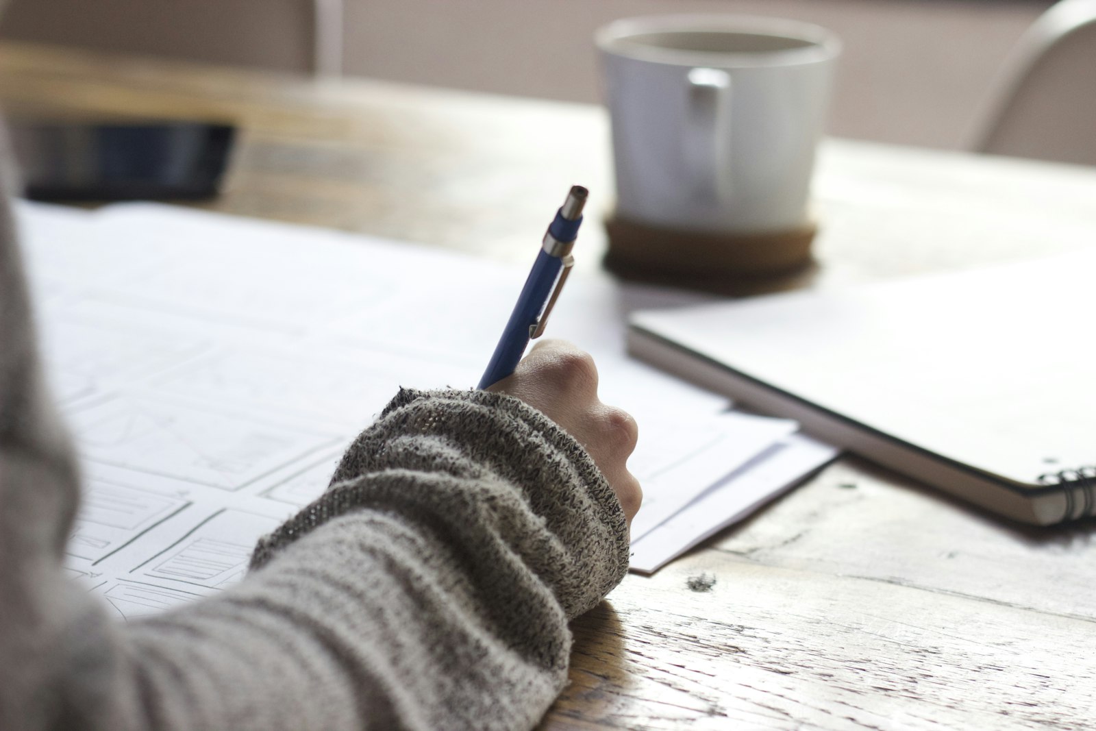 A person writing notes at a desk next to a mug and a tablet